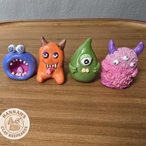 May include: Four small clay monster figurines. The first is a blue round creature with a wide open mouth and spiral eyes. The second is an orange horned monster with sharp teeth and a tongue. The third is a green teardrop-shaped one-eyed creature. The fourth is a textured pink monster with three eyes and two horns.