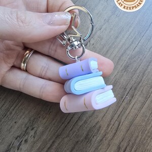 May include: A keychain with three small clay books in pastel shades of lavender, blue, and pink. The books have white pages and gold detailing. The keychain has a silver-coloured clasp and a star-shaped connector. The text "Hannah's Clay Keepsakes" is visible.