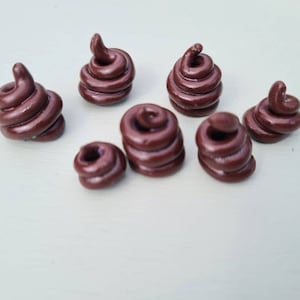May include: Seven small, brown, swirl-shaped decorative objects are displayed. Each object has a rounded base and a spiraling top, resembling a stylized poop emoji. They are arranged on a light-coloured surface.