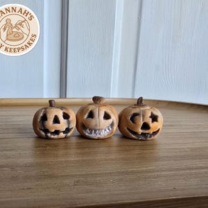 May include: Three small, hand-sculpted clay pumpkins with carved jack-o'-lantern faces, featuring weathered tan and brown tones. Each pumpkin has a distinct expression with cut-out eyes and mouths. A circular logo reads Hannah's Clay Keepsakes.