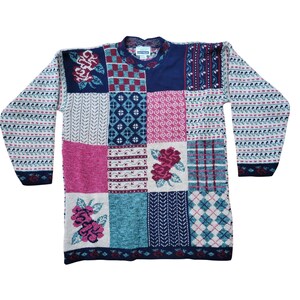 May include: A colourful patchwork sweater with a variety of patterns. The sweater features squares of pink, blue, white, and navy blue, with floral and geometric designs. The sleeves have a repeating pattern of white, pink, and navy blue.