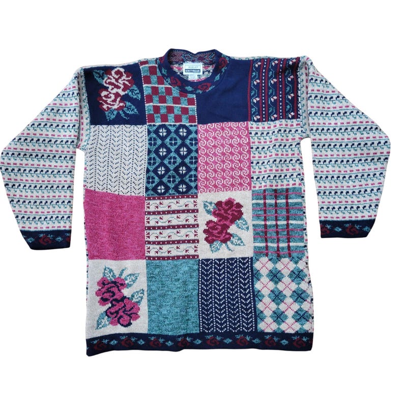 Op de afbeelding: Een patchworktrui met verschillende patronen en kleuren. De trui heeft vierkanten in roze, blauw, wit en marineblauw, met bloemen- en geometrische ontwerpen. De mouwen hebben een herhalend patroon van wit, roze en marineblauw.