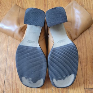 May include: A pair of brown leather knee-high boots with black soles. The boots have a square toe and a block heel. The soles are marked "Leather Upper/Man Made Sole Made in China".