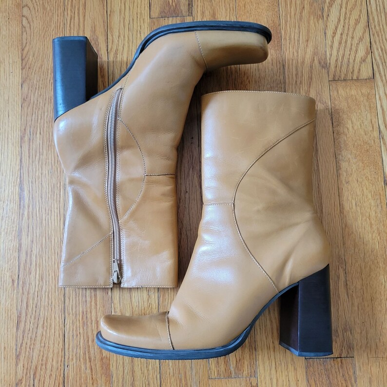 May include: A pair of tan leather knee-high boots with a chunky heel. One boot is unzipped and the other is zipped up.