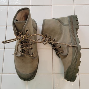 May include: A pair of olive green canvas boots with matching laces. The boots have a high-top design and a rubber sole. The laces are brown with yellow detailing. The boots are on a white tiled surface.