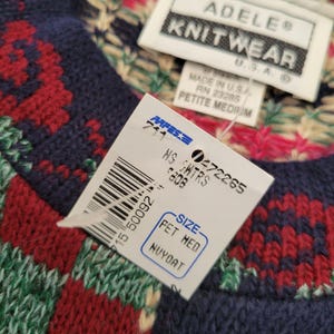 Op de afbeelding: Close-up van een gebreide trui met een label. De trui heeft een patroon van rode, groene en witte ontwerpen op een marineblauwe achtergrond. Het label luidt "ADELE KNITWEAR" en "PETITE MEDIUM".