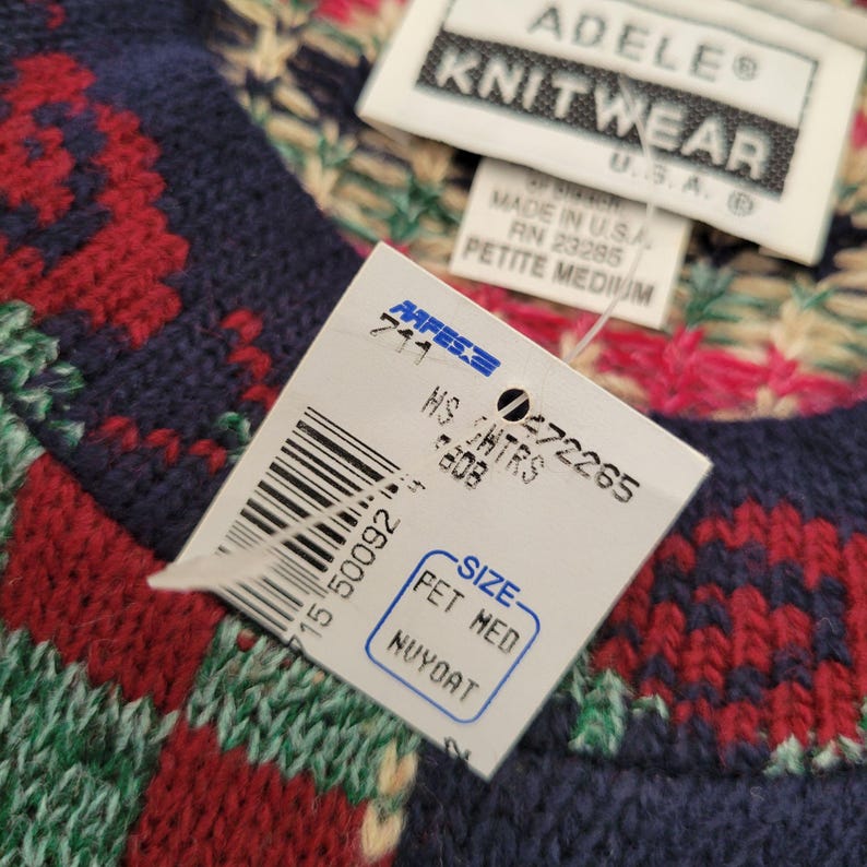 Op de afbeelding: Close-up van een gebreide trui met een label. De trui heeft een patroon van rode, groene en witte ontwerpen op een marineblauwe achtergrond. Het label luidt "ADELE KNITWEAR" en "PETITE MEDIUM".