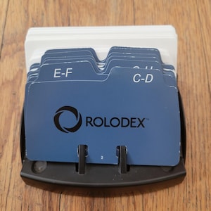 May include: A black plastic Rolodex desktop card file organizer featuring blue alphabetical index divider cards with white text labels including E-F and C-D, along with a stack of blank white contact cards held in a curved base structure.