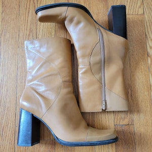 May include: A pair of tan leather heeled boots with a square toe. The boots have a zipper on the inside of each boot and a block heel.