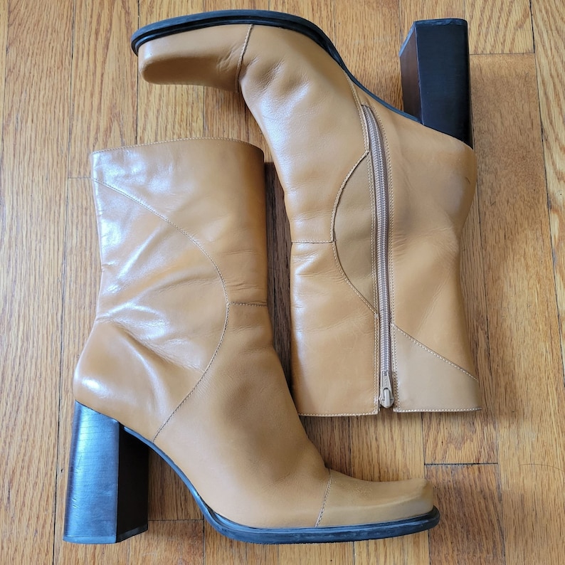 May include: A pair of tan leather heeled boots with a square toe. The boots have a zipper on the inside of each boot and a block heel.