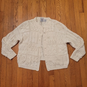 May include: Cream-colored, button-down cardigan sweater with a variety of textured knit patterns. The sweater features a round neckline, long sleeves, and a series of buttons down the front. The knit patterns include floral, geometric, and cable designs.