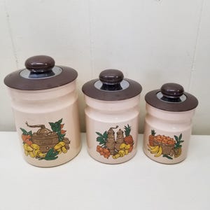 Peut inclure: Ensemble de trois bocaux de cuisine vintage. Les bocaux sont blanc cassé avec des couvercles marron et des accents argentés. Chaque bocal présente un motif différent de fruits et de légumes. Le plus grand bocal a un motif de moulin à café.