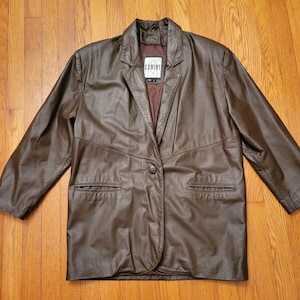 May include: A brown leather blazer with a single-button closure, two front pockets, and a notched lapel. The blazer has a label that reads "COMINT". The jacket is displayed on a wooden surface.