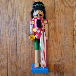 Può includere: Una figura schiaccianoci in legno con capelli neri, una corona di foglie verdi e una camicia rossa con motivi floreali bianchi. Indossa una ghirlanda di fiori gialla, tiene un secchiello giallo e una tavola da surf bianca a righe rosse.