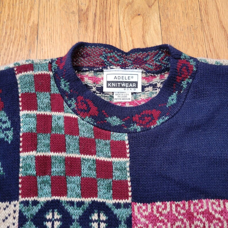 Op de afbeelding: Een vintage Adele Knitwear trui met een patchwork design. De trui heeft een marineblauwe basis met secties van bordeauxrode, teal en cr&egrave;mekleurige patronen. De halslijn en manchetten hebben een patroonrand. Het label luidt "ADELE KNITWEAR USA".