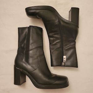 May include: A pair of black leather ankle boots featuring a square toe, a thick block heel, and a platform sole. The boots include a side zipper closure with a silver-toned pull tab and a small red, white, and blue flag detail on the heel.