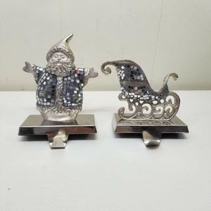 May include: Two silver Christmas stocking holders. One is a Santa Claus figure with a mosaic-style coat. The other is a sleigh with a mosaic-style seat. Both have a rectangular base with a hook for hanging.
