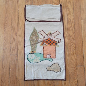 Op de afbeelding: Een rechthoekige, crèmekleurige stoffen wandhanger met een bruine rand. Het ontwerp toont een patchwork windmolen, boom en wolk. De windmolen heeft een oranje dak met een bloemenpatroon, de boom is bruin van textuur. De stijl is rustiek.