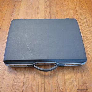 Puede incluir: Un maletín gris oscuro con un borde plateado y un asa negra. El maletín rectangular está cerrado, con una superficie texturizada. El asa está centrada en la parte delantera y la funda parece estar hecha de un material duradero.