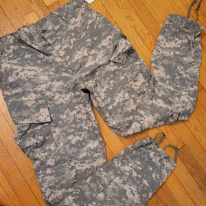 May include: Digital camouflage cargo pants in shades of gray, tan, and green. The pants feature multiple pockets, a button closure, and drawstrings at the ankles. The fabric has a pixelated pattern.