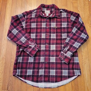 Puede incluir: Una camisa de franela a cuadros rojos y blancos con mangas largas y cierre de botones. La camisa tiene un bolsillo en el pecho izquierdo y el nombre de la marca "Wrangler" es visible en la etiqueta.