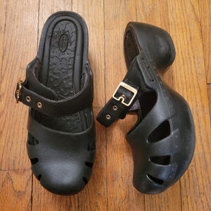 May include: Pair of black clogs with a strap and buckle closure. The clogs have a molded design with cut-out details on the sides. The buckles are gold-colored.