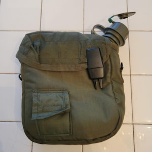 May include: Olive green military canteen with a matching cover. The cover has a small pocket and a flap closure. The canteen has a black plastic clip and a green cap. The letters "U.S." are visible on the cover.