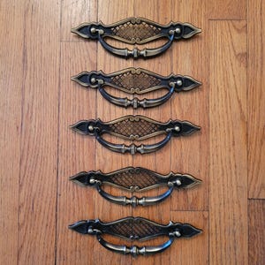 May include: Five antique bronze drawer pulls with a decorative lattice design. The pulls have a curved handle and ornate end pieces. The hardware is set against a wooden background.