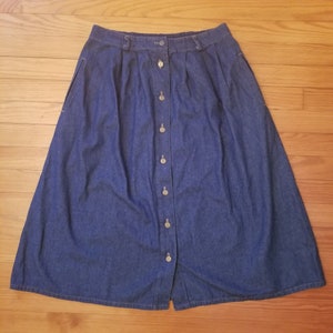 May include: A dark blue denim skirt with a button front and a pleated design. The skirt has pockets on the sides.