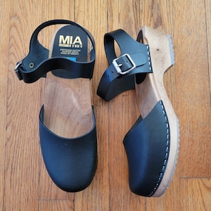 Może przedstawiać: Para czarnych drewniaków ze skórzanymi paskami i drewnianymi podeszwami. Drewniaki mają pasek na kostkę z klamrą. Marka "MIA" jest widoczna na wkładce. Buty leżą na drewnianej powierzchni.
