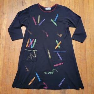 May include: A black long-sleeve dress featuring a crew neckline with a thin red trim and a scattered print of colorful crayons and wavy crayon-drawn lines. The dress includes two front patch pockets, each decorated with crayon graphics.