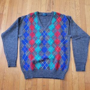 May include: A gray V-neck sweater with a colorful argyle pattern. The argyle design features red, blue, and teal diamonds, with yellow accents. The sweater has long sleeves and ribbed cuffs and hem.