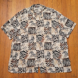 May include: Short-sleeved button-down shirt with a repeating pattern of zebras and cheetahs in a grid design. The shirt features a collared neckline and a beige background with black and brown animal prints.