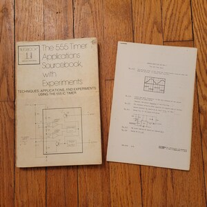 Pode incluir: Um livro aberto intitulado "The 555 Timer Applications Sourcebook, with Experiments" e uma folha de papel solta. A capa do livro tem um diagrama esquemático e o texto "TECHNIQUES, APPLICATIONS, AND EXPERIMENTS USING THE 555 IC TIMER".