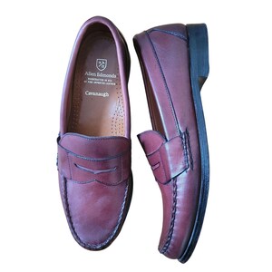 May include: A pair of burgundy leather Allen Edmonds Cavanaugh penny loafers. The shoes have a classic design with a stitched vamp, a penny keeper strap, and a black sole. The brand name and style are visible on the insole.
