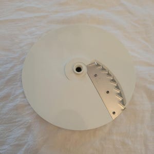 May include: A white, circular food processor blade with a stainless steel slicing blade attached. The blade has a central hub with a hole and is set against a white background.