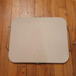 May include: A rectangular, off-white baking stone with rounded corners and two metal handles. The stone is set on a wooden surface.