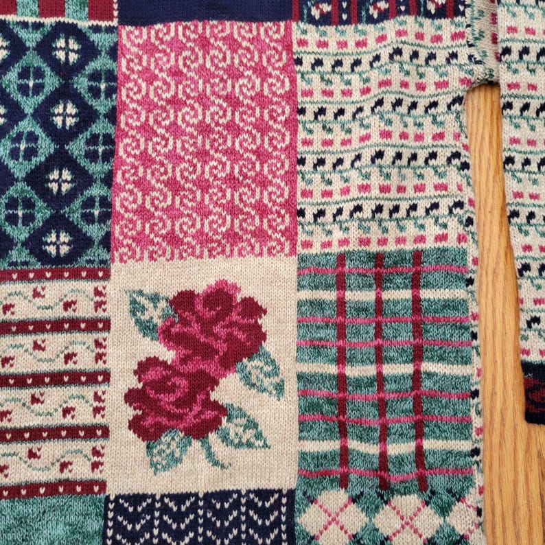 Op de afbeelding: Close-up van een patchwork trui met verschillende breipatronen. De trui heeft vierkanten in teal, marine, cr&egrave;me en roze. E&eacute;n vierkant heeft een paar rode rozen. Andere vierkanten hebben geometrische en bloemmotieven.