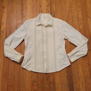 May include: Cream-colored long-sleeve button-down shirt. The shirt features vertical embroidered panels down the front and on the cuffs. The collar and cuffs are detailed with a subtle textured design.