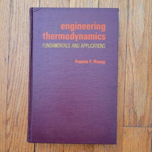 May include: A hardcover book titled "engineering thermodynamics" with the subtitle "FUNDAMENTALS AND APPLICATIONS" and the author's name, Francis F. Huang, in orange text on a purple cover. The book is on a wooden surface.