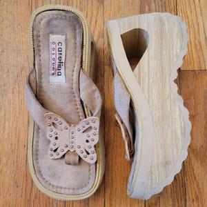 Op de afbeelding: Een paar beige suède platformsandalen met een hoge houten sleehak. De teenslipperband is voorzien van een decoratieve vlinderuitsnede versierd met kleine zilveren studs. Een wit label op het voetbed toont de tekst Carolina Colours.