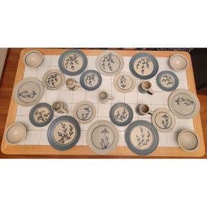 Puede incluir: Una colección de vajilla de cerámica, que incluye platos, cuencos y tazas, dispuesta sobre una mesa de madera. Los platos presentan un diseño floral azul y blanco. Las tazas y los cuencos son de color crema liso.