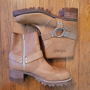 Könnte beinhalten: Ein Paar hellbraune Durango-Leder-Ankle Boots. Die Stiefel haben einen seitlichen Reißverschluss, einen Riemen mit silberner Hardware und eine klobige Profilsohle. Das Wort "DURANGO" ist auf der Seite eines Stiefels geprägt.