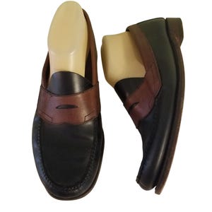 May include: Pair of black leather penny loafers with brown leather saddle straps. The shoes are displayed on shoe forms against a white background. The soles are brown.