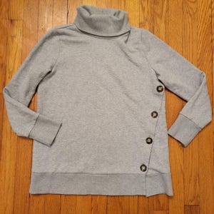 Op de afbeelding: Een lichtgrijs sweatshirt met een coltrui en lange mouwen. Het sweatshirt heeft drie grote, decoratieve knopen aan de linkerkant. De zoom en manchetten zijn een donkerdere grijstint.