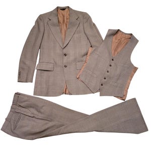 Puede incluir: Un traje de tres piezas con estampado de cuadros en color marrón claro, compuesto por una americana de dos botones con solapas de muesca, un chaleco de cinco botones y un pantalón. Las prendas tienen forro interior beige y botones.