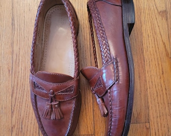 May include: A pair of brown leather loafers with braided trim and tassel details. The shoes have a classic design with a rounded toe and a low heel. The loafers are displayed on a wooden surface.
