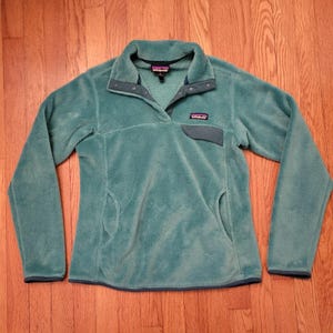 May include: A teal fleece pullover with a button-up collar and a small chest pocket. The long-sleeved top has two side pockets and a dark teal trim. A small brand logo is visible on the chest.