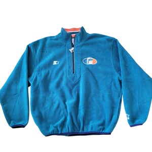 Pode incluir: Pulôver de lã azul-petróleo com fecho de zíper de um quarto. Apresenta o logotipo do Miami Dolphins no peito e um logotipo Starter na manga e no peito. A gola tem acabamento laranja e branco.
