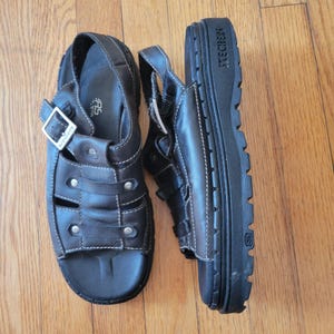 May include: A pair of black leather sandals with a buckle closure and a thick platform sole. The sandals have a woven design and the brand name "TECHNIC" is printed on the sole.